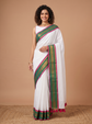 A woman modeling a white saree with a green and magenta border, paired with a white sleeveless blouse, set against a solid beige studio background.