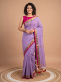 A woman models a lavender purple silk saree with a magenta pink border and gold zari detailing, paired with a matching pink blouse, standing on a circular woven mat against a solid beige backdrop.