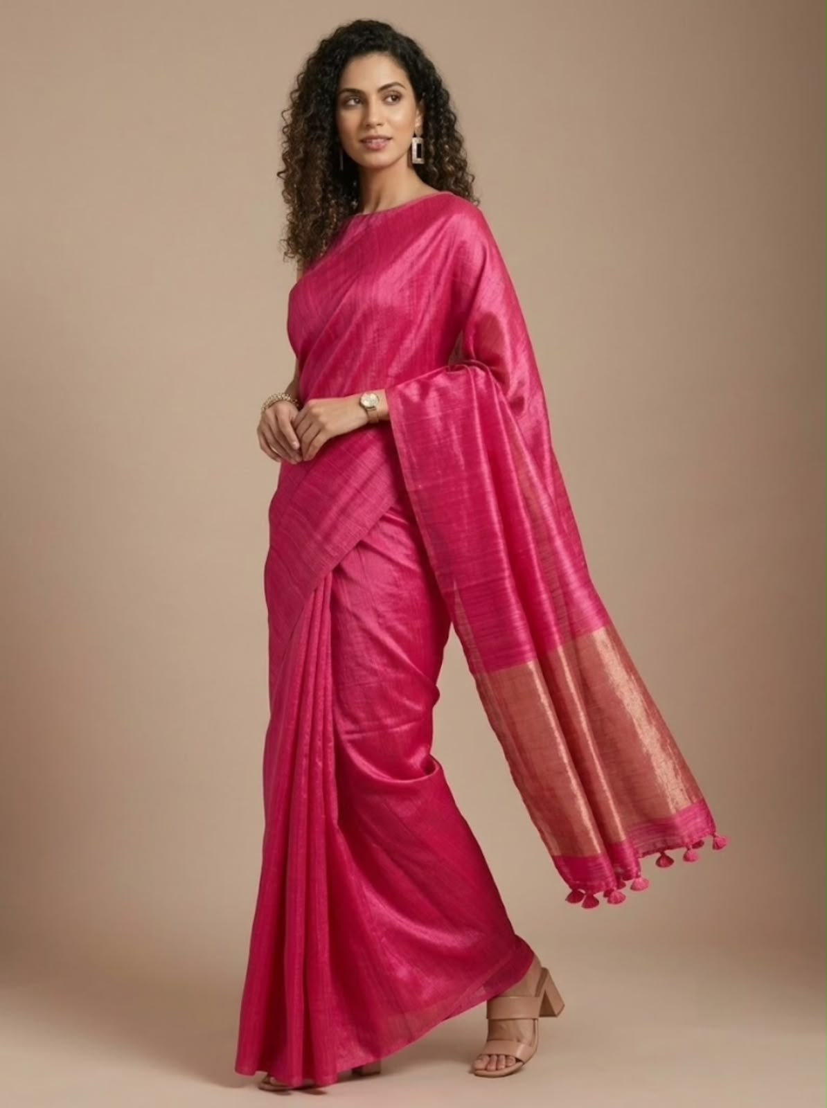 A woman modeling a bright pink silk saree with a metallic gold border and matching pink tassel trim, posed against a solid beige studio background.