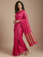 A woman modeling a bright pink silk saree with a metallic gold border and matching pink tassel trim, posed against a solid beige studio background.