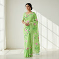 A woman models a light green saree with pink floral and green vine embroidery, standing in a bright room with soft window shadows.