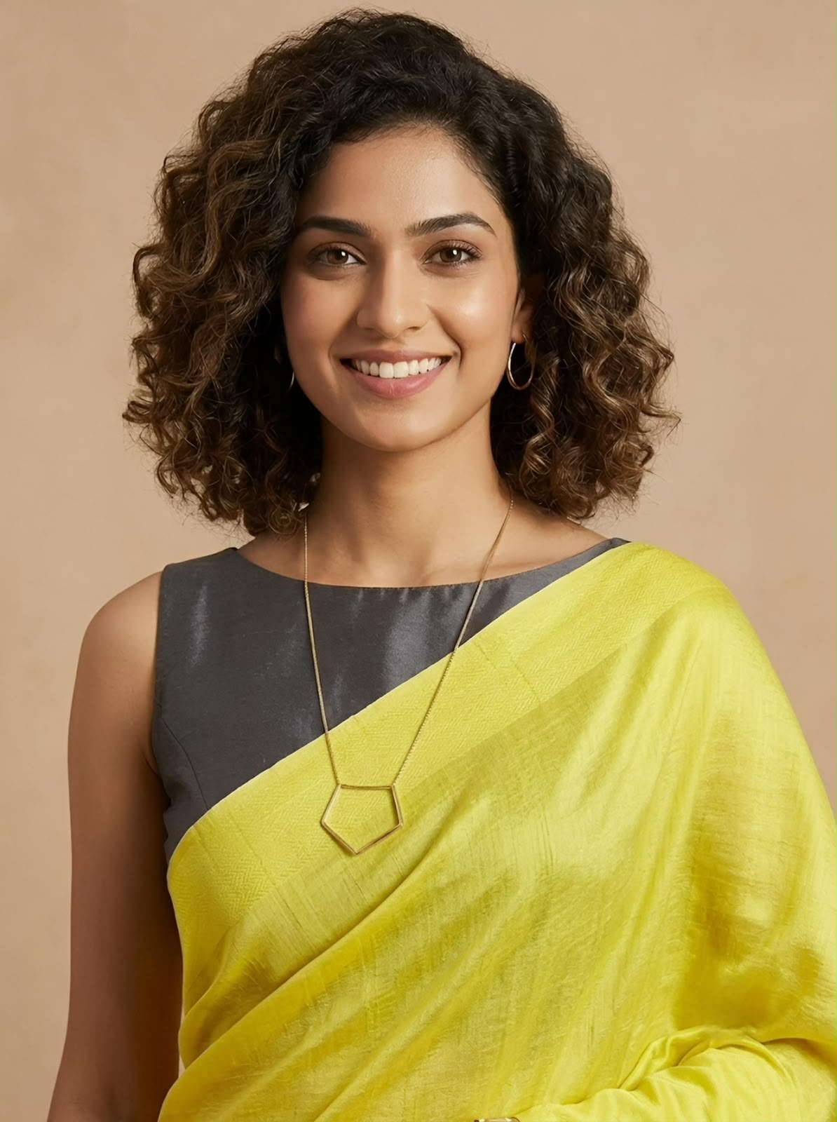 A woman with curly brown hair wearing a bright yellow and dark gray draped sari with a gold pendant necklace, set against a solid beige background.