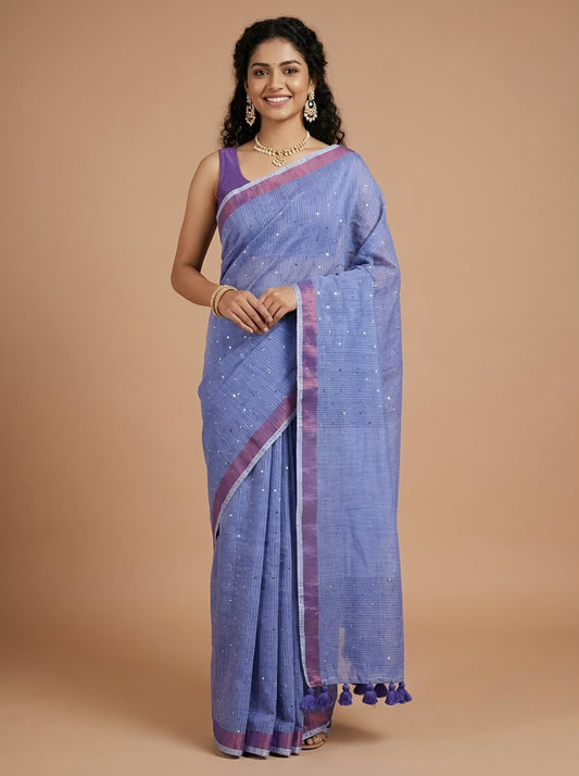 A woman modeling a lavender-blue saree with purple border accents and small silver embellishments, standing against a solid beige studio background.