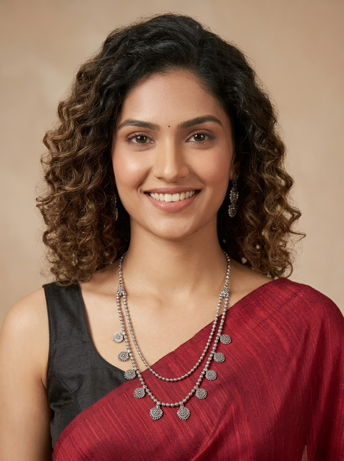 A smiling woman with curly brown hair wearing a dark gray sleeveless top and a deep red sari, accessorized with a silver-toned layered necklace and earrings, set against a warm beige background.