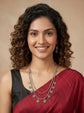 A smiling woman with curly brown hair wearing a dark gray sleeveless top and a deep red sari, accessorized with a silver-toned layered necklace and earrings, set against a warm beige background.