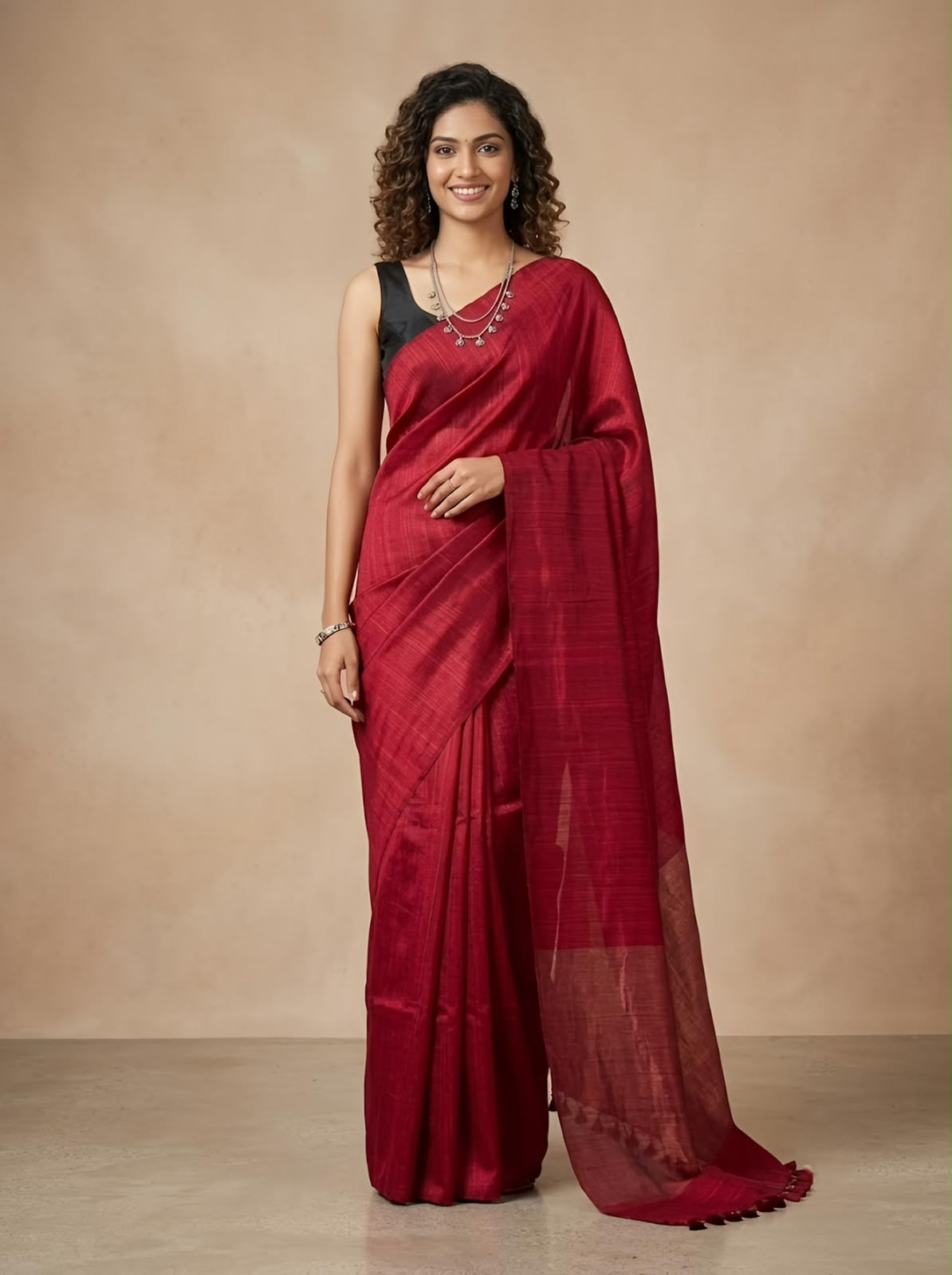 A woman modeling a deep red silk saree with a black sleeveless blouse, standing against a warm beige studio backdrop.