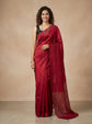 A woman modeling a deep red silk saree with a black sleeveless blouse, standing against a warm beige studio backdrop.