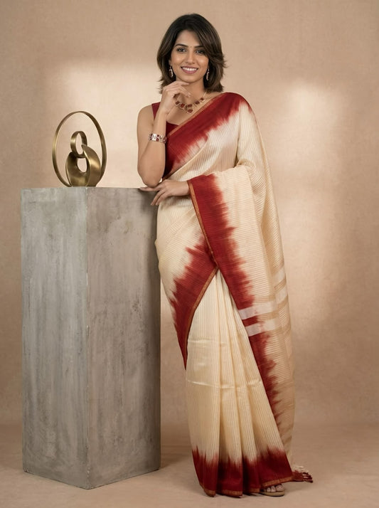 A woman wearing a cream-colored silk saree with a deep red tie-dye border and pallu, standing next to a gray concrete pedestal with a gold sculpture against a beige background.