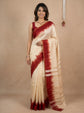 A woman modeling a cream-colored silk saree with a deep red tie-dye border and matching red sleeveless blouse, set against a warm beige textured studio backdrop.