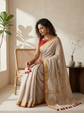 A woman seated on a rattan chair wearing a light beige saree with a red blouse, gold border, and red tassel accents, in a softly lit indoor setting with a plant and side table.