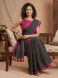 A woman models a black and gray checkered saree with a vibrant magenta pink border and blouse, seated on a traditional wooden bench in a warm, decorated room.