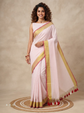 A woman models a light pink saree with a gold and red border, paired with a matching sleeveless blouse, standing on a round woven mat against a neutral beige background.