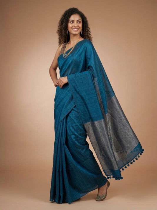 A woman modeling a teal blue saree with a wide charcoal gray border and matching blue tassel trim, posed against a solid warm beige studio background.