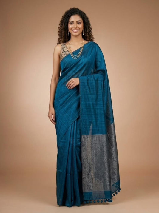 A woman modeling a teal blue saree with a silver woven border and a matching blue blouse, standing against a solid beige studio background.