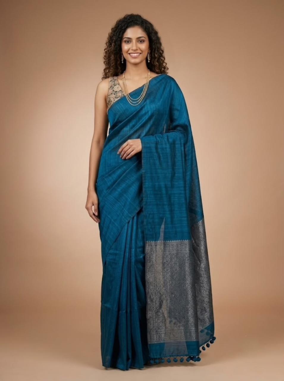 A woman modeling a teal blue saree with a silver woven border and a matching blue blouse, standing against a solid beige studio background.