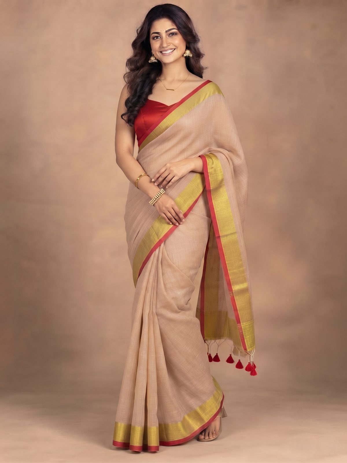 A woman modeling a beige silk saree with a red and gold border, paired with a red blouse, standing against a warm beige studio backdrop.