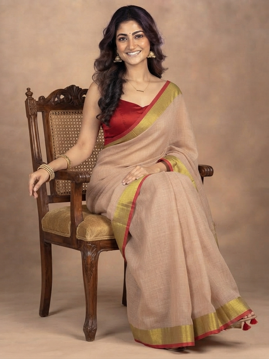 A woman seated on a carved wooden chair wearing a beige saree with a red blouse and gold and red border details, posed against a warm beige studio backdrop.