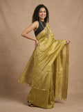 A woman modeling a golden-yellow silk saree with a black sleeveless blouse, standing against a textured beige studio backdrop.