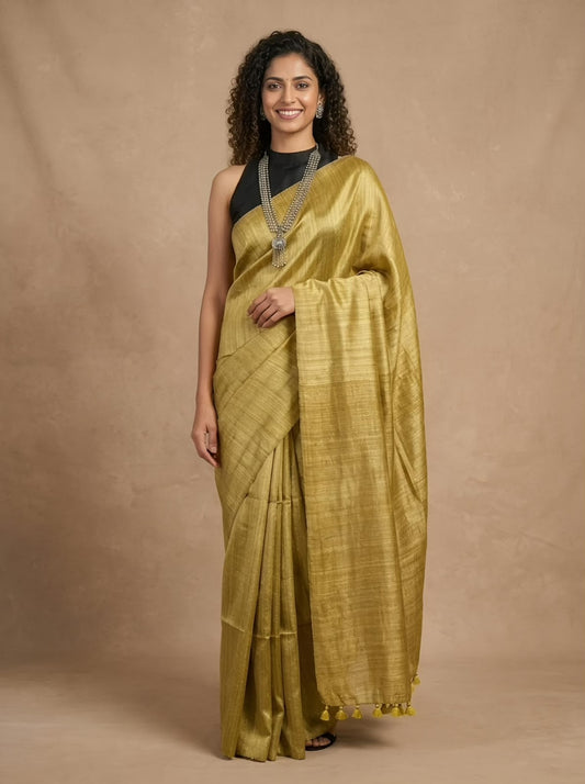 A woman modeling a golden-yellow silk saree with a black sleeveless top, standing against a textured beige studio backdrop.