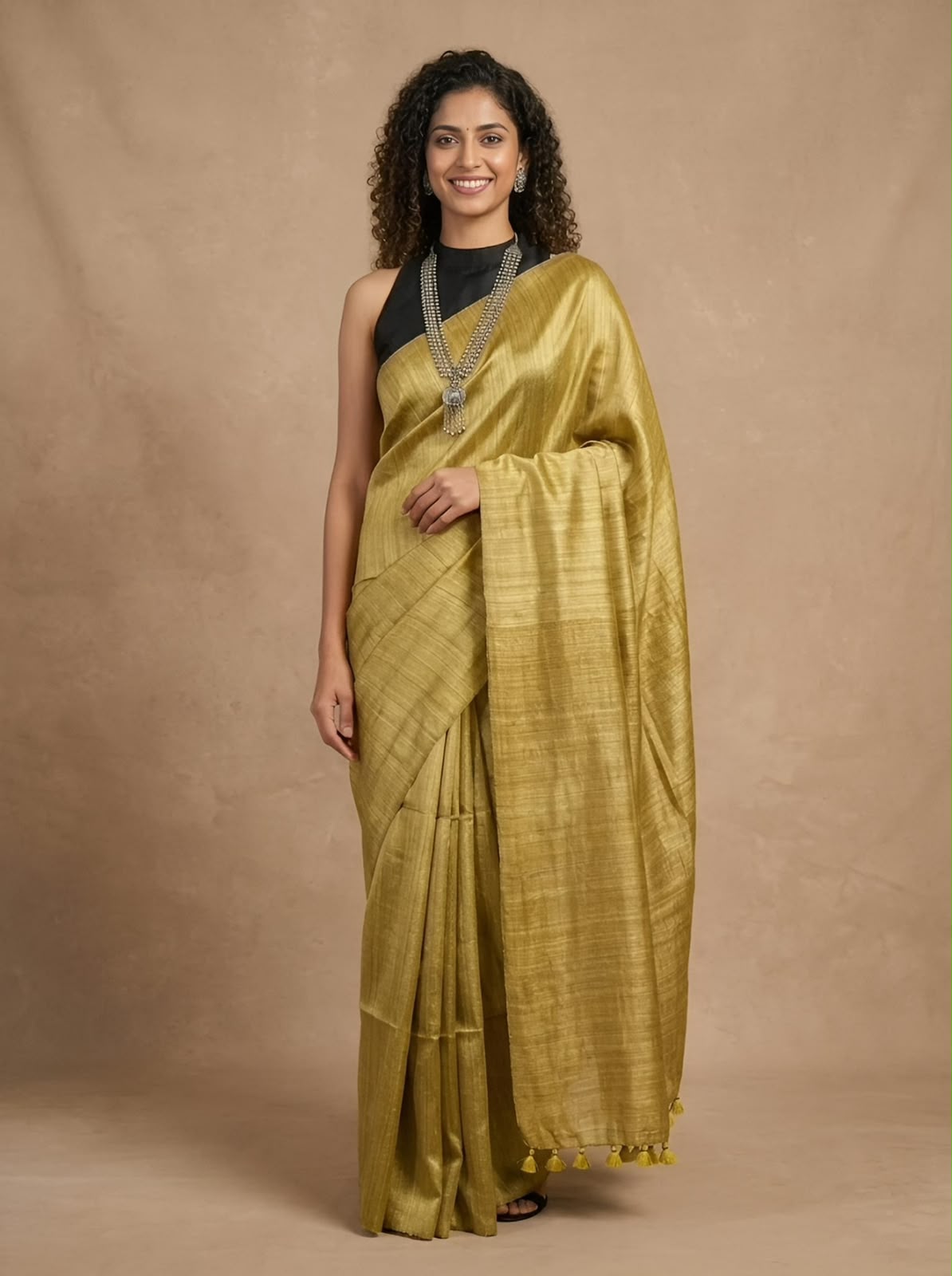 A woman modeling a golden-yellow silk saree with a black sleeveless top, standing against a textured beige studio backdrop.