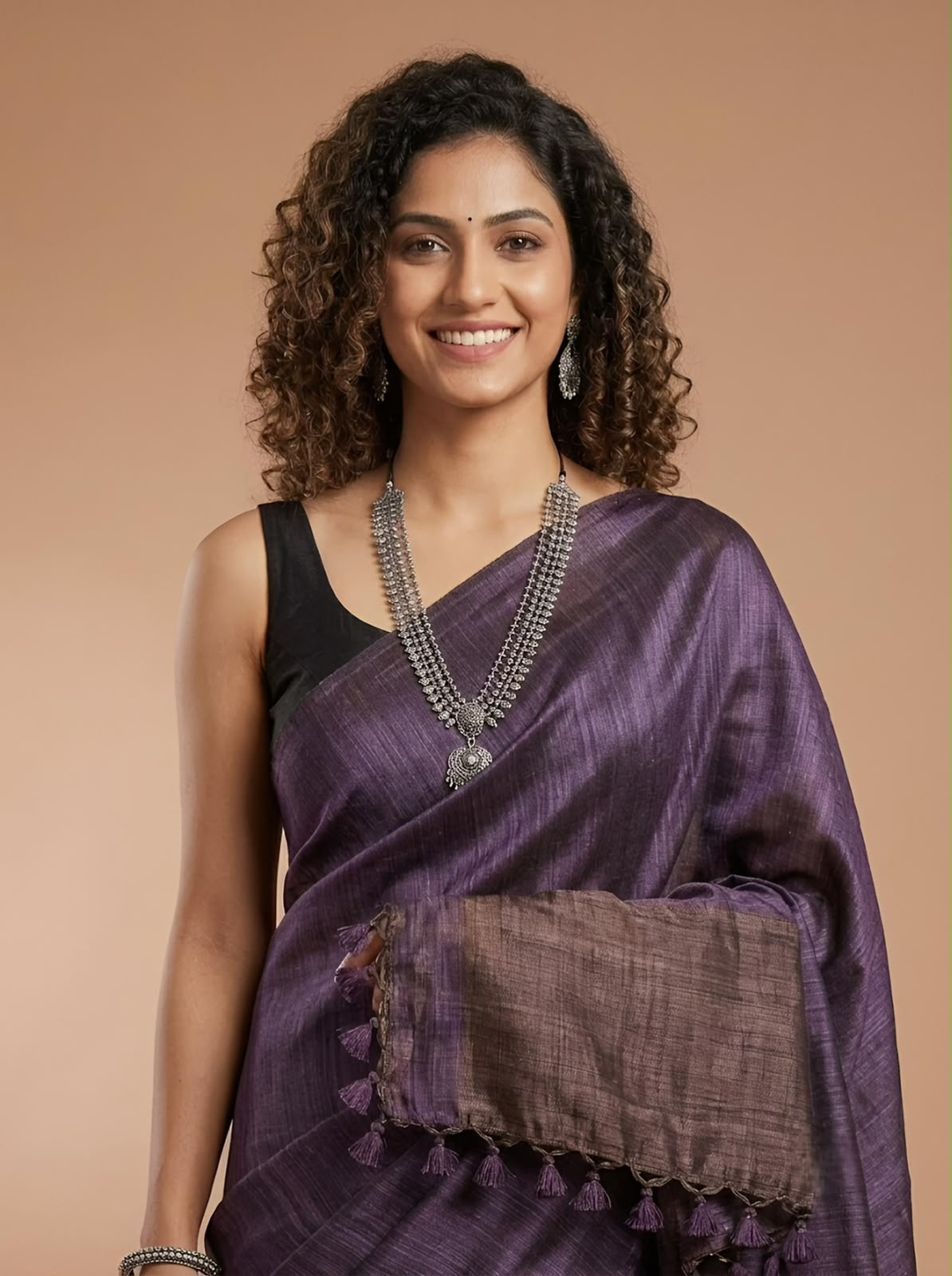 A woman with curly dark hair models a purple silk saree with a textured brown pallu and a black sleeveless blouse, accessorized with silver jewelry, against a solid beige background.