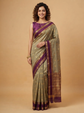 A woman modeling a traditional Indian saree in olive green with a deep purple border and blouse, set against a solid beige studio background.