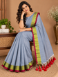 A woman seated indoors wearing a light blue saree with a lime green and magenta border and red tassels, posed in a warm neutral room with a bench, plants, and a vase.