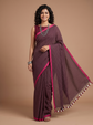 A woman modeling a deep maroon purple cotton saree with a fine white checkered pattern, bright pink borders, and a matching pink blouse, set against a solid tan studio background.