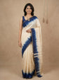 A woman modeling a cream-colored cotton saree with a deep blue tie-dye pattern and a matching blue blouse, standing against a neutral beige backdrop.