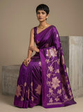 A woman models a purple silk saree with metallic gold floral patterns, seated on a concrete block against a neutral beige background.