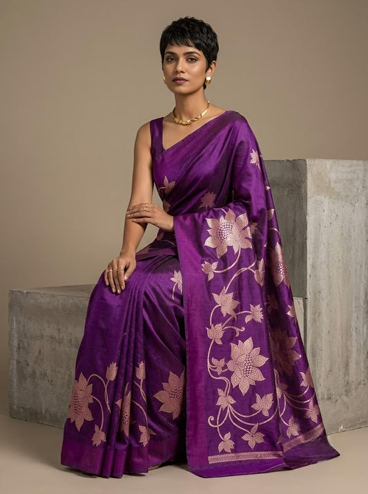 A woman models a purple silk saree with metallic gold floral patterns, seated on a concrete block against a neutral beige background.